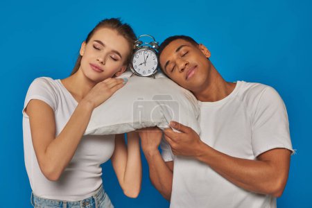 positive interracial couple sleeping together on pillow near alarm clock on blue background