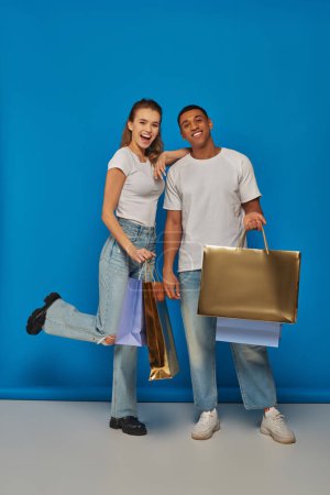 holiday sales, happy interracial couple  holding shopping bags and standing on blue backdrop