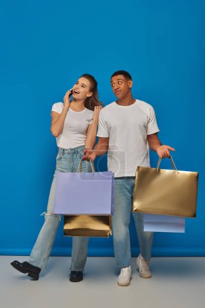 consumerism, interracial couple  holding shopping bags on blue backdrop, woman talking on smartphone