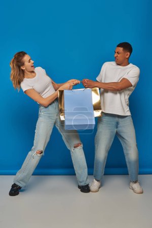 consumerism, interracial man and woman pulling shopping bags on blue backdrop, holiday sales