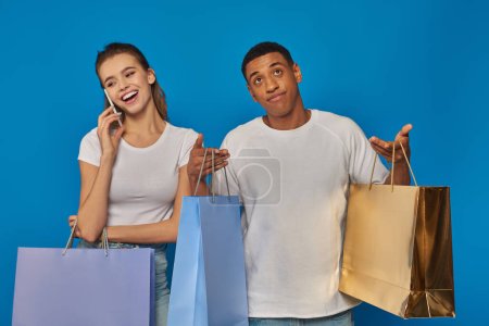 consumerism, happy woman having phone call while african american boyfriend holding shopping bags