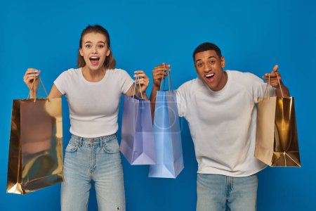 excited interracial couple holding shopping bags and looking at camera on blue backdrop, consumerism
