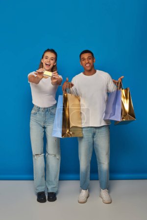 alegre pareja interracial sosteniendo bolsas de compras y tarjeta de crédito sobre fondo azul, consumismo