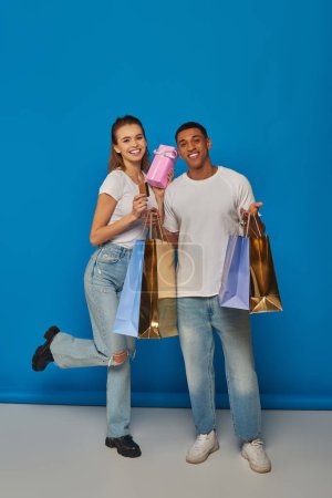 Foto de Feliz pareja multicultural sosteniendo bolsas de compras y tarjeta de crédito sobre fondo azul, regalo de compra - Imagen libre de derechos