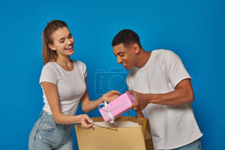 happy multicultural couple putting present into shopping bag on blue backdrop, consumerism concept