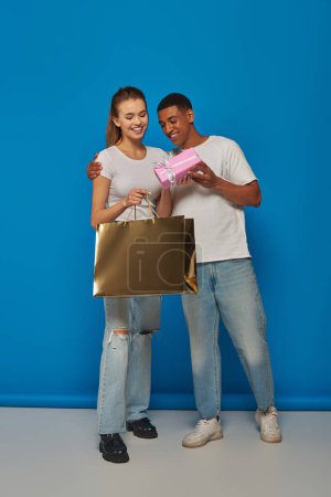 cheerful interracial couple buying present, holding shopping bag on blue backdrop, consumerism