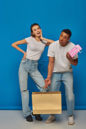 positive multicultural couple buying present, holding shopping bag on blue backdrop, consumerism