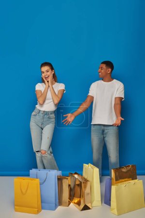 Diverses Paar, afrikanisch-amerikanischer Mann zeigt auf Einkaufstüten neben aufgeregter Frau vor blauem Hintergrund