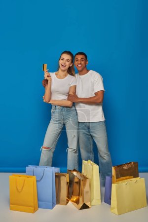 positive multicultural couple standing near shopping bags and holding credit card on blue backdrop