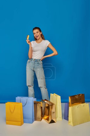 mujer feliz celebración de la tarjeta de crédito y de pie cerca de bolsas de compras sobre fondo azul, longitud completa