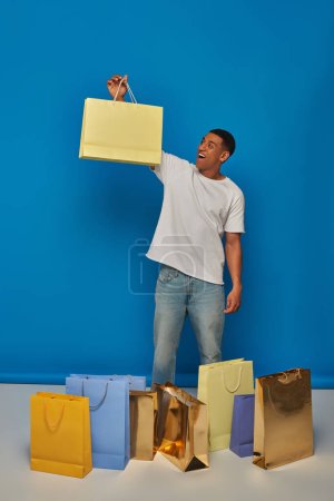 hombre afroamericano emocionado en traje casual sosteniendo bolsas de compras en el telón de fondo azul, compra de juerga