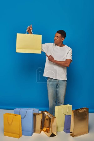 hombre afroamericano feliz en traje casual apuntando a la bolsa de compras en el telón de fondo azul, compra de juerga
