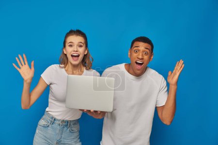 excitada pareja multicultural gesticulando cerca del ordenador portátil sobre fondo azul, concepto de trabajo remoto