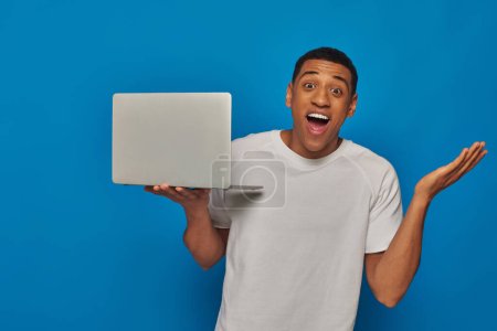 Téléchargez les photos : Excité homme afro-américain gestuelle et tenant ordinateur portable sur fond bleu, concept de travail à distance - en image libre de droit