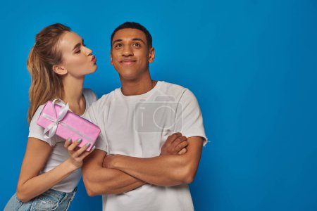 pretty woman holding present near cheerful african american man on blue backdrop, air kiss