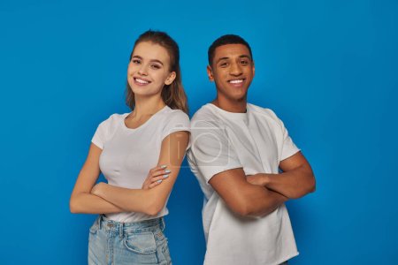 cheerful multicultural couple standing with folded arms and looking at camera on blue background