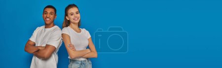 Photo for Happy interracial couple standing with folded arms and looking at camera on blue background, banner - Royalty Free Image