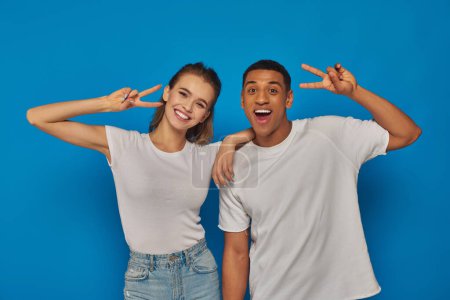 happy multicultural couple showing peace sign and looking at camera on blue background, positivity
