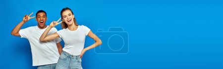 Photo for Cheerful multicultural couple showing v sign and looking at camera on blue background, banner - Royalty Free Image