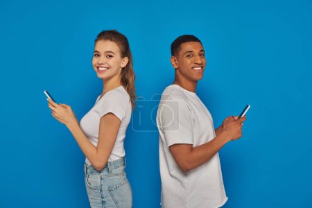 Foto de Alegre pareja multicultural de pie espalda con espalda y el uso de teléfonos inteligentes sobre fondo azul - Imagen libre de derechos