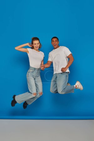 positive multicultural couple holding hands and jumping  on blue background, full length view