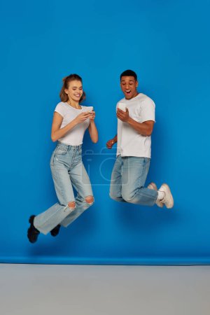 glückliches multikulturelles Paar mit Smartphones und Springen auf blauem Hintergrund, Ansicht in voller Länge