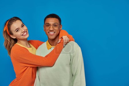 happy woman in orange cardigan hugging african american man in eyeglasses on blue backdrop, style