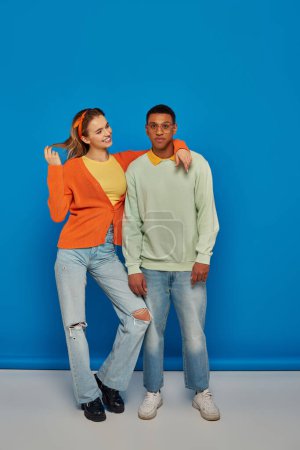 interracial couple in casual attire on blue backdrop, young woman hugging african american man