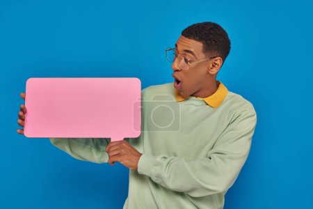 shocked african american man in eyeglasses looking at blank speech bubble on blue backdrop Poster 671417072