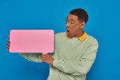 shocked african american man in eyeglasses looking at blank speech bubble on blue backdrop Poster #671417072
