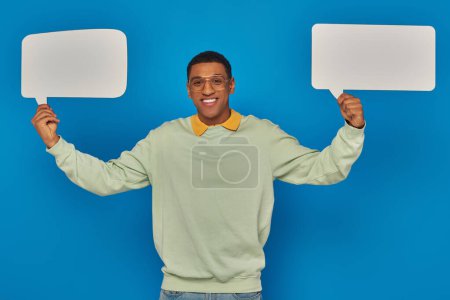 cheerful african american man in eyeglasses and smart casual clothes holding blank speech bubbles