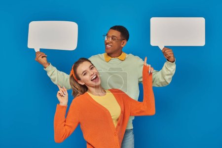 cheerful diverse couple in smart casual clothes holding blank speech bubbles on blue backdrop