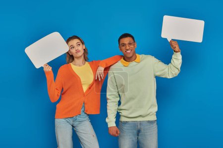 cheerful interracial couple in smart casual clothes holding blank speech bubbles on blue backdrop