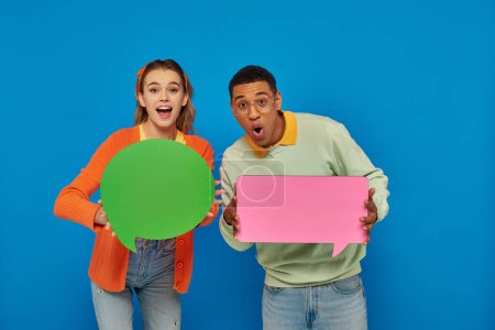 Photo for Excited interracial couple in smart casual clothes holding blank speech bubbles on blue backdrop - Royalty Free Image