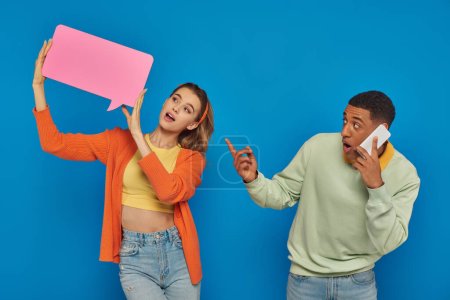 amazed african american man in glasses pointing at woman with speech buttle on blue background