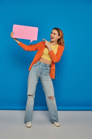 Photo for Excited woman in casual attire pointing at speech bubble on blue background, space for text - Royalty Free Image