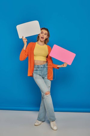 Photo for Astonished young woman in casual attire holding speech bubbles on blue background, space for text - Royalty Free Image