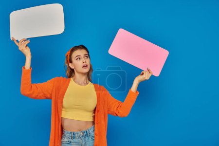 surprised young woman in casual attire holding speech bubbles on blue background, space for text