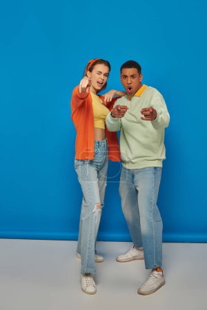 excited multicultural couple looking at camera and pointing with fingers on blue backdrop