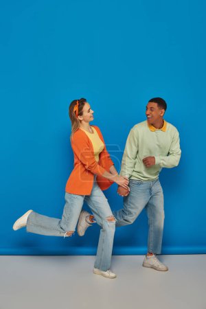 glückliches multikulturelles Paar in lässiger Kleidung, Händchen haltend und auf blauem Hintergrund laufend, lächelnd