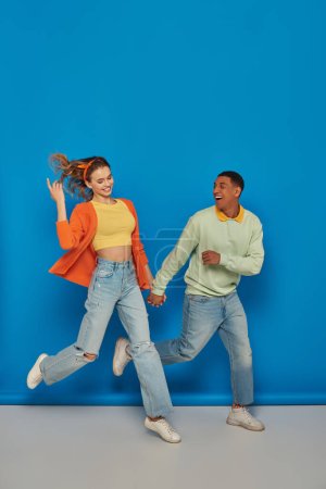 Photo for Cheerful multicultural couple in casual attire holding hands and running on blue background, smile - Royalty Free Image