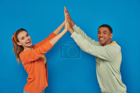excitada pareja multicultural en atuendo casual dando alta cinco y sonriendo sobre fondo azul