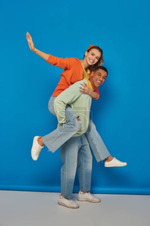 happy african american man piggybacking cheerful girlfriend on blue background, having fun