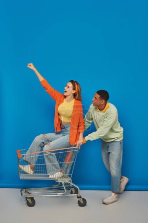 hombre afroamericano positivo montando carrito de compras con novia dentro de ella sobre fondo azul