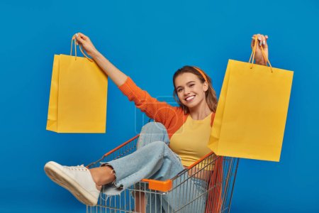 positive junge Frau sitzt im Einkaufswagen und hält Einkaufstüten auf blauem Hintergrund, Kaufrausch