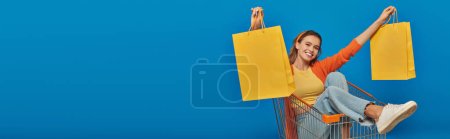 happy woman sitting in cart and holding shopping bags on blue background, buying spree, banner