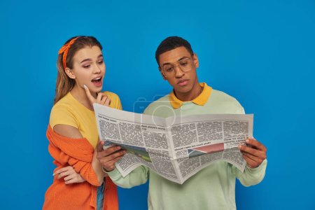 Photo for Amazed woman reading newspaper near calm african american man in eyeglasses on blue backdrop - Royalty Free Image