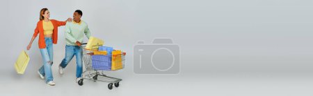 excited interracial couple walking with trolley and shopping bags on grey backdrop, banner