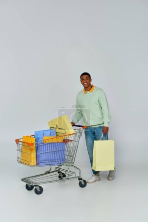 excited african american man standing with trolley and shopping bags on grey backdrop, consumerism