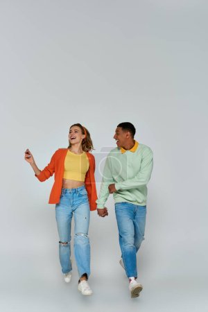 glückliches interrassisches Paar in lässiger Kleidung, Händchen haltend und vor grauem Hintergrund zusammen laufend